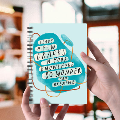 Spiral blue 6” x 8” notebook with abstract shapes and a hand-lettered inspirational quote on the cover about wonder. 