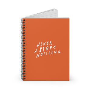 Spiral notebook, standing up on table, showing the orange cover with a big white quote about noticing the little things. 