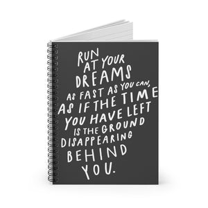 Spiral notebook, standing up on table, showing the cover with a big black and white quote about following your dreams. 