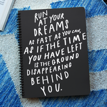 Load image into Gallery viewer, Black spiral journal with white hand-lettered quote about following your dreams, on blue and white blanket.
