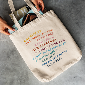 Woman’s hands pulling fashion magazines out of trendy cotton tote bag with a quote about creativity printed on the front.