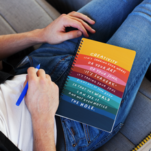 Load image into Gallery viewer,  Man reclining on floor, holding a pen, with colourful spiral journal on his lap featuring a creativity quote on cover.
