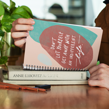 Load image into Gallery viewer, Woman at her desk, reading her open spiral journal on top of a pile of books. Journal features inspiring quote art on cover.
