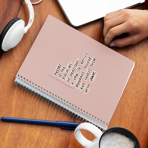 Pink spiral journal on desk, with poetry quote on cover, alongside a mug, pen, headphones, and laptop. 