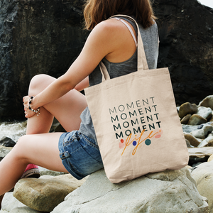 Woman sitting on rocks with her tote bag over her shoulder. Tote bag features quote about mindfulness.